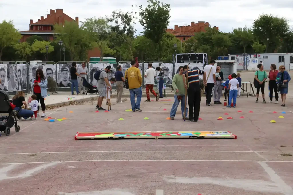 Fotorgafía de un evento de juegos tradicionales con un grupo de personas jugando y fotografías de retratos de gran tamaño en blanco y negro al fondo.